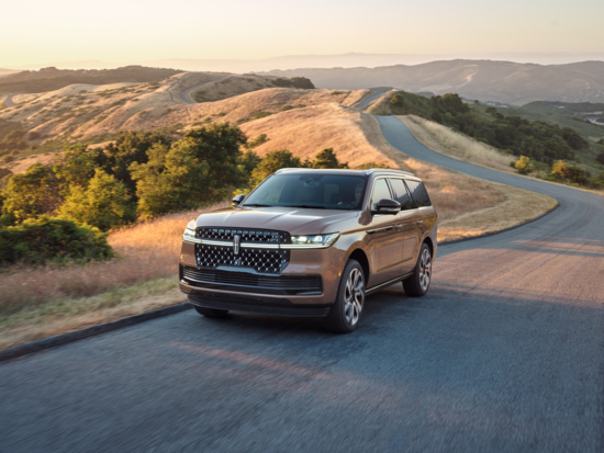 A bronze SUV drives on a winding road through a hilly landscape at sunset. The sky is clear, casting a warm glow over the dry grasses and lush trees.