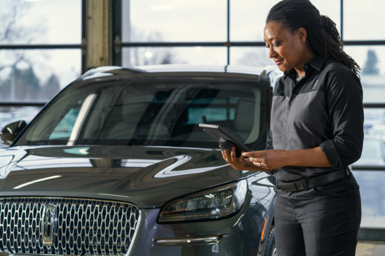 A woman in a service uniform stands next to a luxury SUV in a garage, holding a tablet. She appears focused and professional, with large windows behind her.