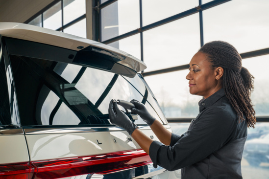 Woman in gloves carefully attaches a logo to the back of a shiny, white SUV inside a garage. The setting conveys focus and precision.