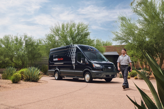 A black mobile service van parked on a sunny, desert road. A man in a uniform walks beside the van, surrounded by green bushes and a clear blue sky.