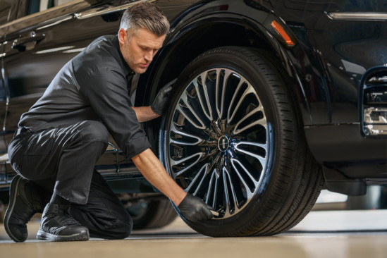 A mechanic in black uniform and gloves inspects a car's polished wheel in a garage, kneeling with focus and precision, highlighting attention to detail.