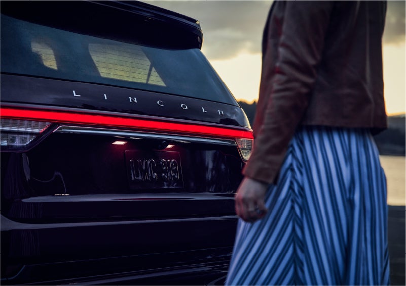 A person is shown near the rear of a 2023 Lincoln Aviator® SUV as the Lincoln Embrace illuminates the rear lights | Korum Lincoln in Puyallup WA