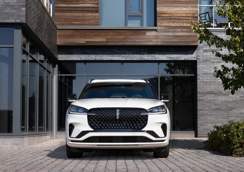 A white 2025 Lincoln Aviator® SUV is shown parked outside a modern building with gray brick and wood accents. | Korum Lincoln in Puyallup WA