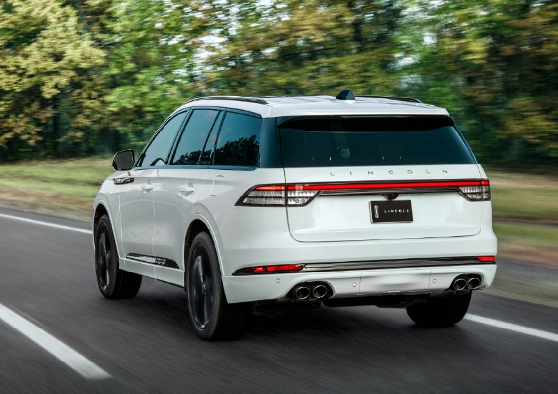 A white 2026 Lincoln Aviator® SUV is being driven away down a road lined with colorful trees. | Korum Lincoln in Puyallup WA